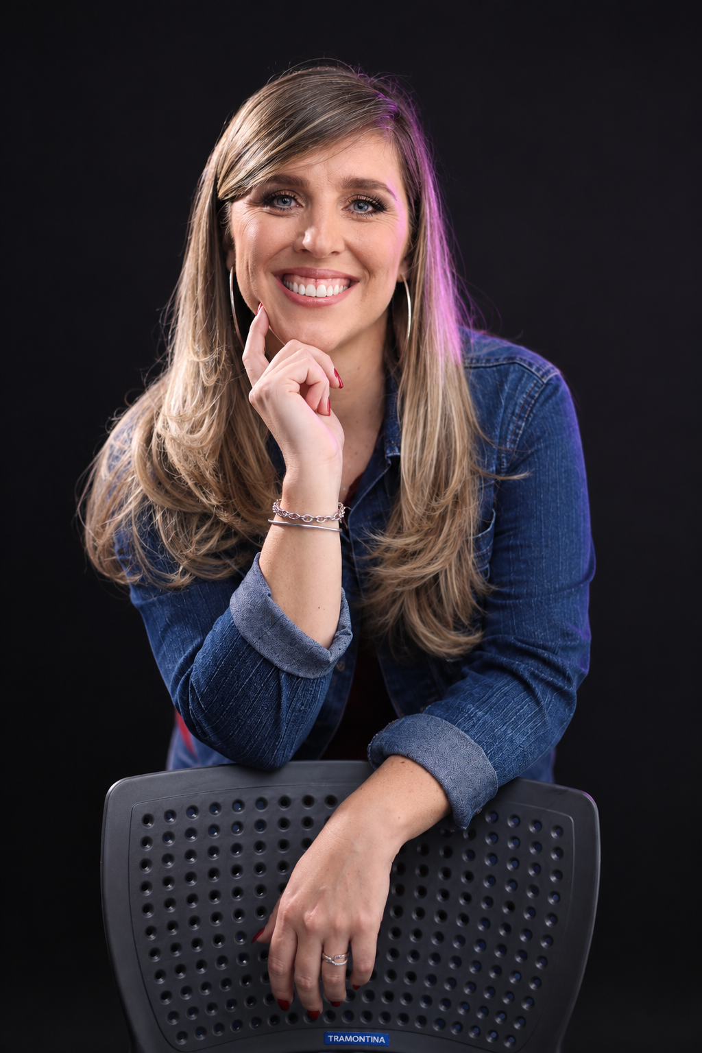 Maureen sorrindo, apoiada em uma cadeira, com camisa jeans.